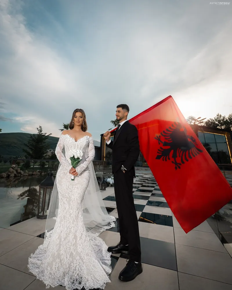 Bride in a lace gown beside the groom holding a red Albanian flag