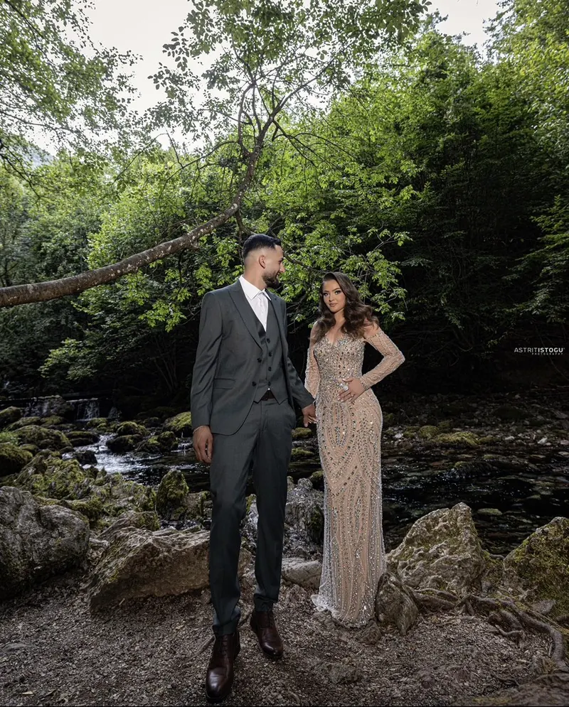 A couple in formal wear standing on mossy rocks by a forest stream
