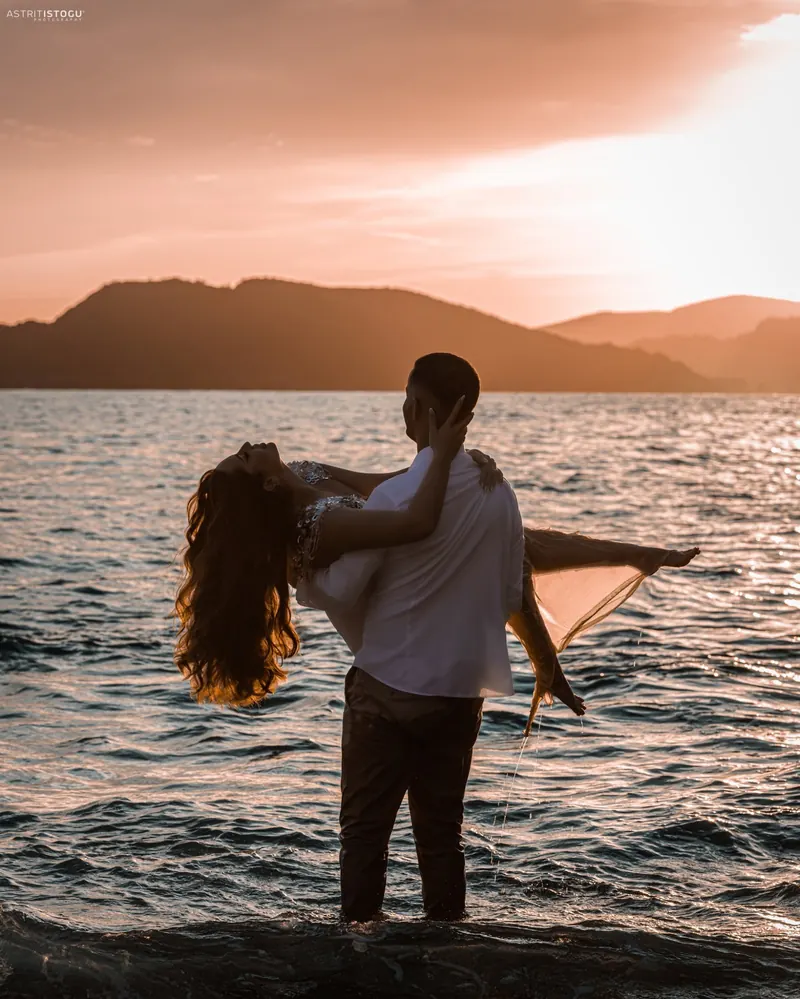 A man carrying his partner through the water at sunset, mountains behind