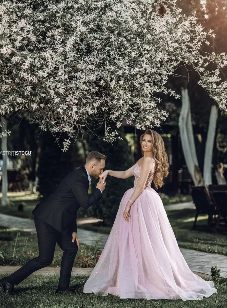A man kneeling to kiss the hand of a woman in a pink gown in a garden