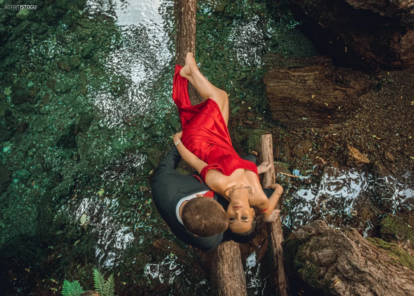 Overhead portrait of a woman in a red dress lying across a fallen log above a forest stream