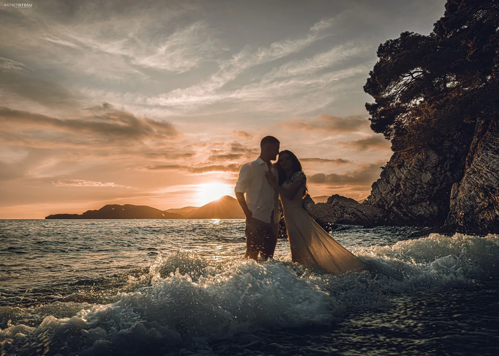 A couple embracing in the surf at sunset beside a rocky coast