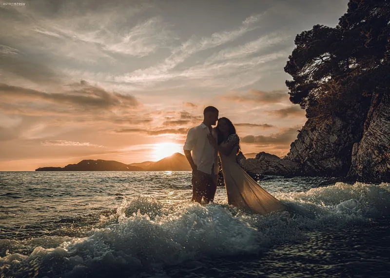 A couple embracing in the surf at sunset beside a rocky coast